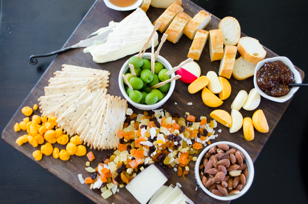 Overhead shot of a square-shaped cutting board filled with various cheeses, nuts, olives and crackers. 