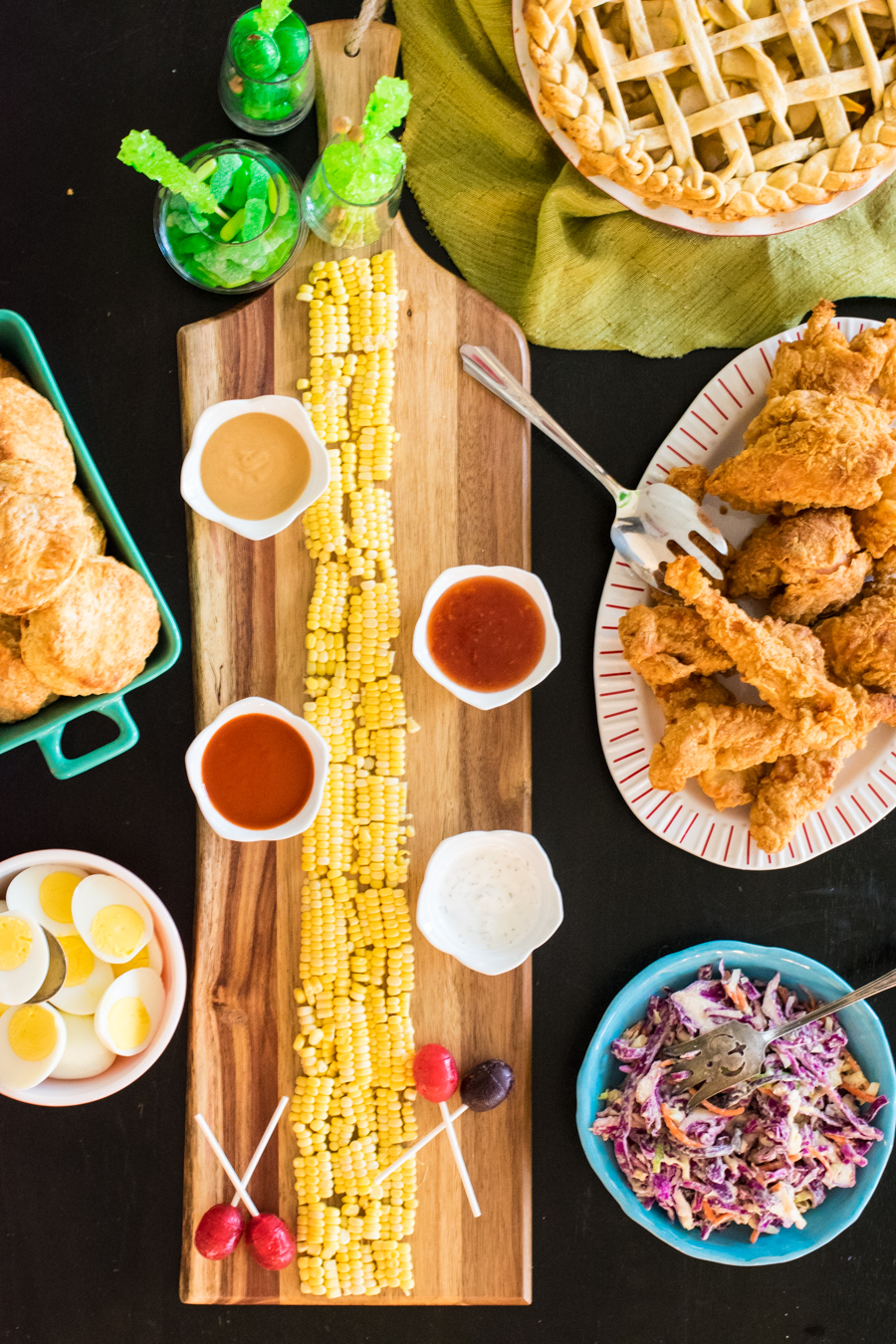 Overhead shot of a table full of food for a Wizard of Oz dinner menu. Yellow Brick Road made of corn kernels in the center, surrounded by lollipops, fried chicken, dips, cole slaw, hard boiled eggs, and baking powder biscuits
