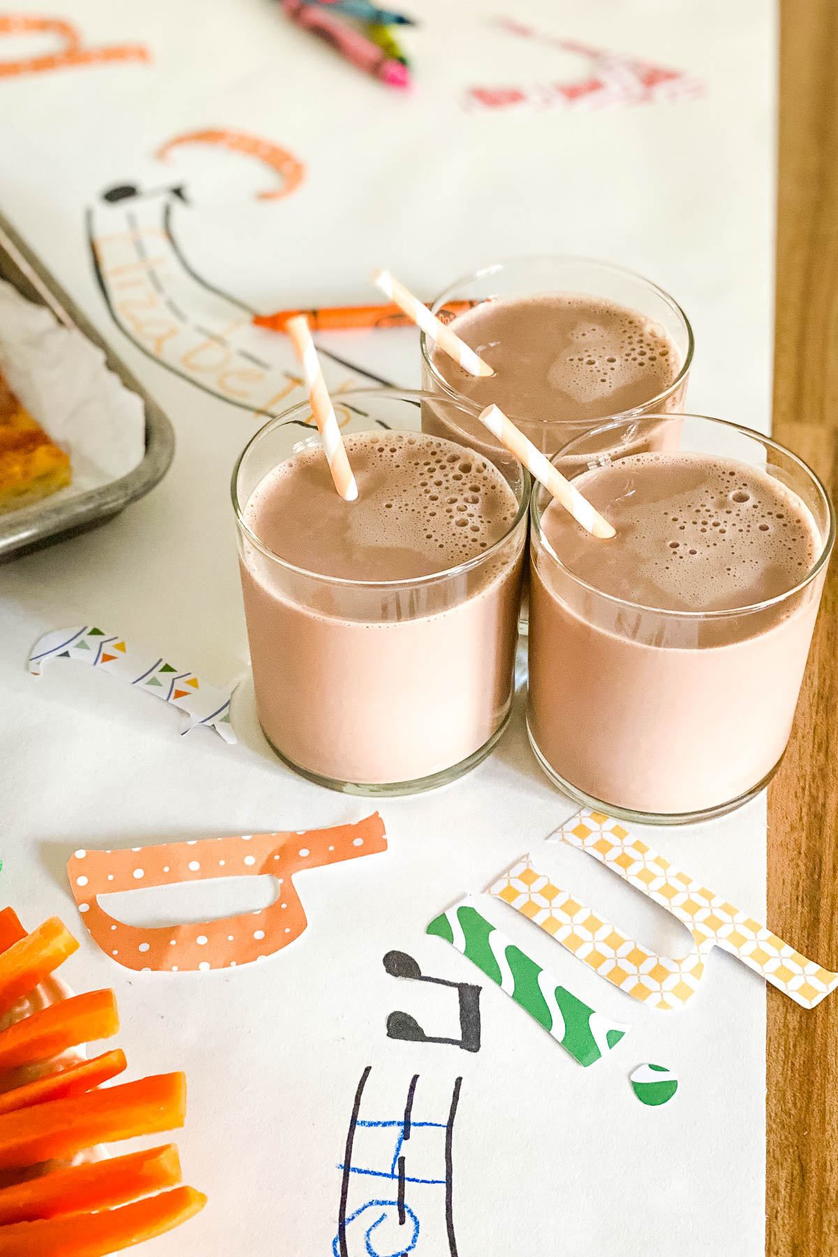 Three short glasses of chocolate milk are in focus in the foreground. They are set on a white paper table runner decorated with colorful paper letters.